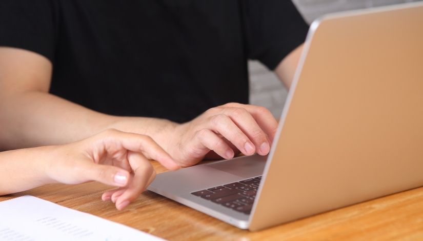 Imagem de um notebook sobre uma mesa com a mão de uma pessoa que veste camiseta preta. Ao seu lado aparece a mão de outra pessoa.
