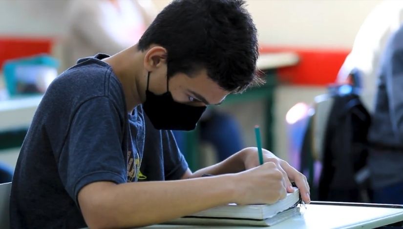 Fotografia de um estudante escrevendo em seu caderno. Ele usa máscara de proteção individual.