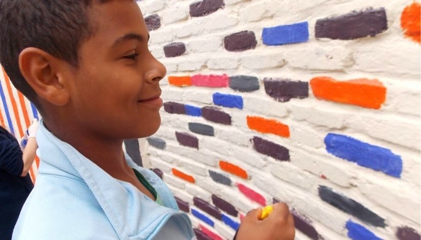 Fotografia de um menino sorrindo. Ele segura um giz amarelo na mão direita e está de frente para uma parede branca com alguns tijolinhos coloridos nas cores laranja, grafite, azul, rosa.