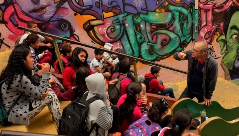 Imagem mostra professor e alunos em uma escadaria durante aula aberta. Ao fundo, um muro grafitado com palavras e cores