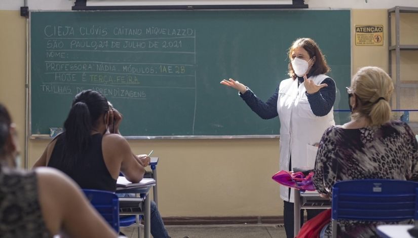 Fotografia de uma professora em momento de aula com estudantes que estão sentadas em suas cadeias. Ao fundo, tem uma lousa verde onde se-lê 