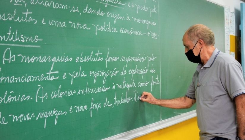 Fotografia de um professor usando máscara de proteção individual. Ele está escrevendo na lousa verde com giz branco.