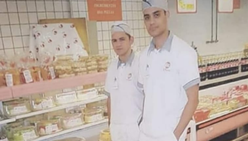 Fotografia de duas pessoas vestidas com o uniforma da padaria. Eles usam toca e estão parados em frente as prateleiras com bolos e pães.
