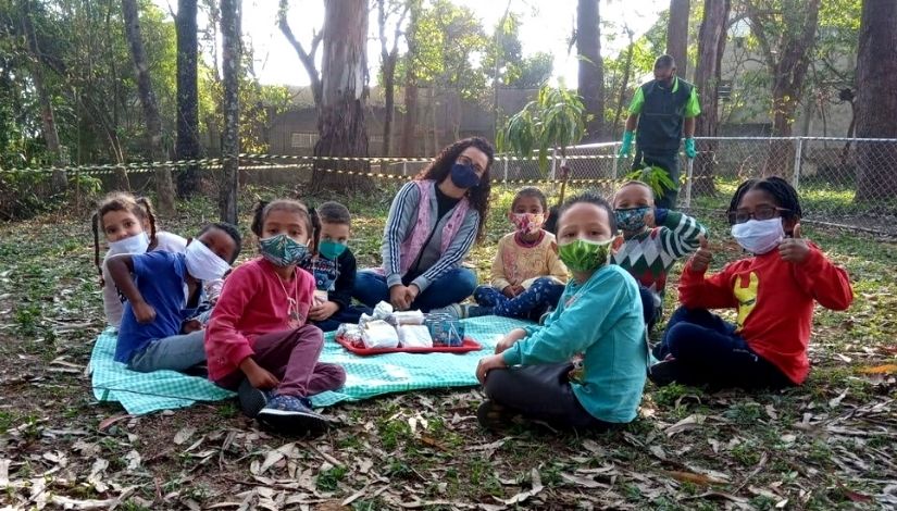 Fotografia de uma professora e seus estudantes da EMEI sentados sobre uma toalha xadrez verde. Eles estão em um piquenique na área externa com grama, folhas secas e árvores. Ao fundo um homem está em pé, ele usa roupas e luvas verdes.