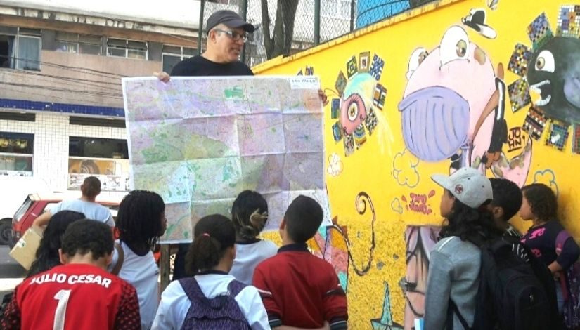 Imagem mostra professor com um mapa nas mãos explicando conteúdo para os alunos. Grupo está em um local aberto e há uma parece amarela com desenhos ao fundo