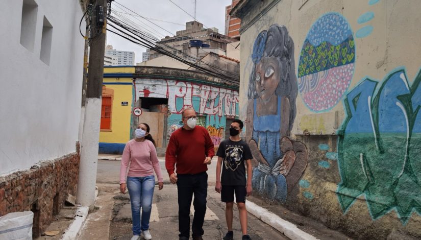 Professor Paulo veste blusa vermelha e caminha entre dois alunos sendo uma menina com blusa rosa e calça jeans à sua direita e um menino de camiseta e bermuda preta à esquerda. Eles andam em um corredor estreito do bairro da Liberdade olhando para os imóveis. Uma das casas tem paredes brancas e a outra colorida com a pintura de uma menina negra. 