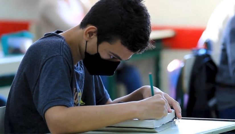 Estudante veste camiseta azul escura, máscara preta e está escrevendo com um lápis verde em um caderno.