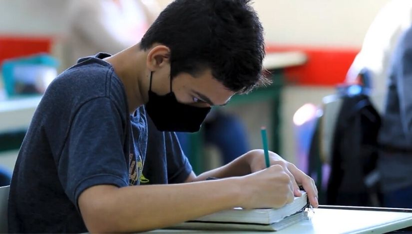 Estudante de camiseta azul, máscara preta, está escrevendo com um lápis em um caderno.
