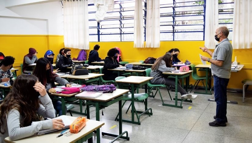 Professor dando aula para uma sala com estudantes. Todos usam máscara de proteção individual.