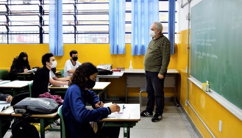 Imagem mostra professor em uma sala de aula com paredes amarelas, em frente a lousa. Os alunos estão em carteiras, copiando os conteúdos do quadro.