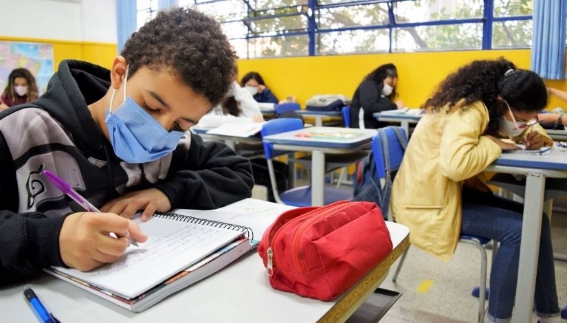 Imagem mostra em primeiro plano um aluno usando moletom preto e cinza, com máscara azul. À sua frente, um caderno e um estojo vermelho. Ao fundo, outros alunos também aparecem escrevendo em cadernos