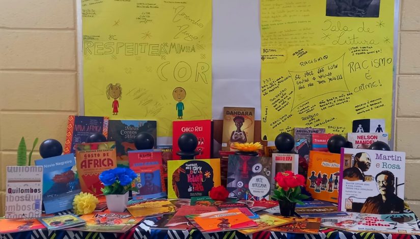Imagem mostra uma mesa com dezenas de livros posicionados. Na parte de trás, cartazes amarelos com frases escritas como "Respeita a minha cor" e "Racismo é crime", escritas pelos estudantes
