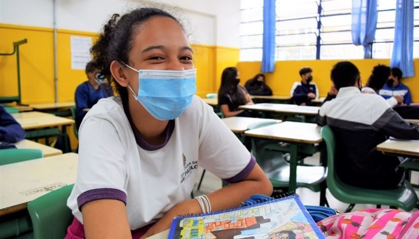 Imagem mostra uma menina em uma sala de aula com a parede amarela ao fundo. Ela usa uma máscara azul, camiseta branca com a logomarca da Prefeitura de São Paulo. Está olhando e sorrindo para a câmera.