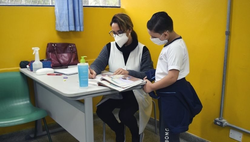 fotografia de professora atendendo um garoto em sua mesa. Todos usam máscara facial.