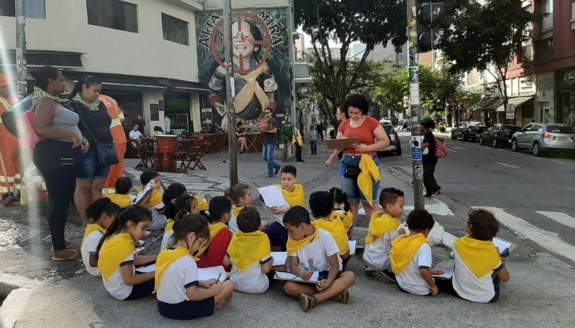 Crianças da Educação Infantil sentadas em uma calçada da capital paulistana acompanhadas da professora e de familiares. As crianças seguram pranchetas sobre o seu colo e lápis, elas estão observando a cidade.