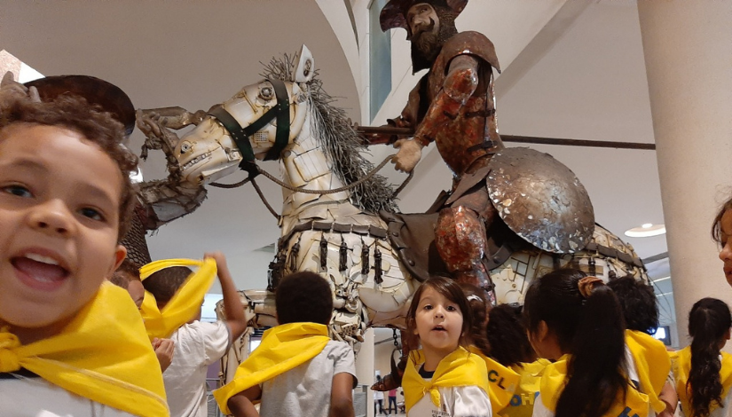 Crianças da Educação Infantil com lencinhos amarelos no pescoço visitando uma exposição com a escultura metálica de Dom Quixote.