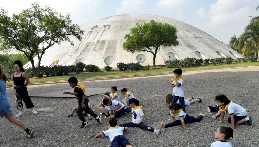 Crianças da Educação Infantil em um espaço aberto sentadas no chão em frente a OCA, no Parque do Ibirapuera.