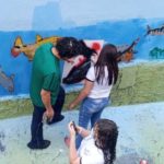 Professor e estudantes fazendo a pintura do mural na parede nos fundos da EMEF. Eles usam a técnica de estêncil.