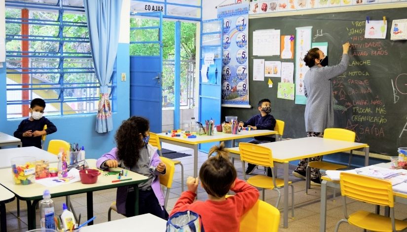 Imagem de uma professora escrevendo na lousa e estudantes pequenos sentados às suas mesas. Todos usam máscaras de proteção individual.