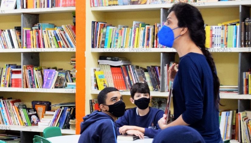 foto de professora na sala de leitura, dois estudantes olham atentamento para ela. Todos usam máscaras