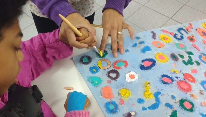 fotografia mostra uma mão infantil sendo auxiliada pela mão de um adulto