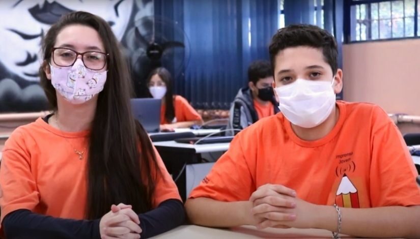 Fotografia de dois estudantes do Projeto Imprensa Jovem. Uma menina e um menino, os dois usam camiseta laranja do Imprensa Jovem e máscara de proteção individual.
