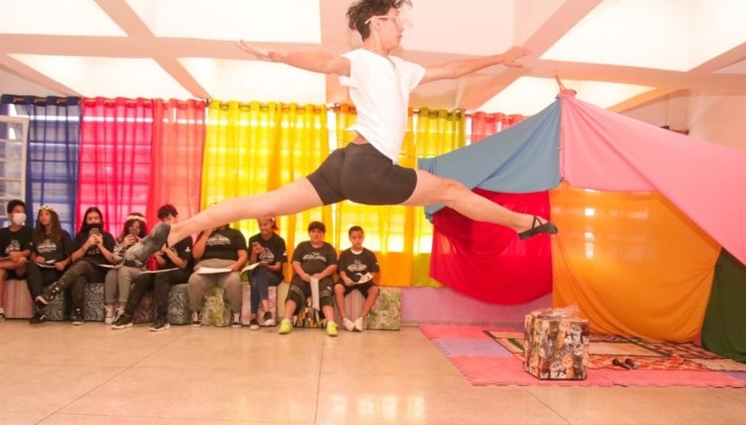 Ao centro, em primeiro plano, dançarino saltando com as pernas e braços esticados. Ao fundo, crianças e adolescentes sentados em puff. Do lado direito, também ao fundo, uma tenda com panos coloridos e tapete.