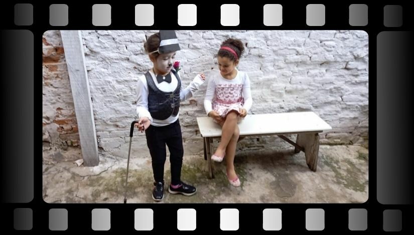 Fotografia de uma criança em pé segurando uma bengala e outra sentada em um banco com as pernas cruzadas. A imagem está dentro de uma moldura de filme.