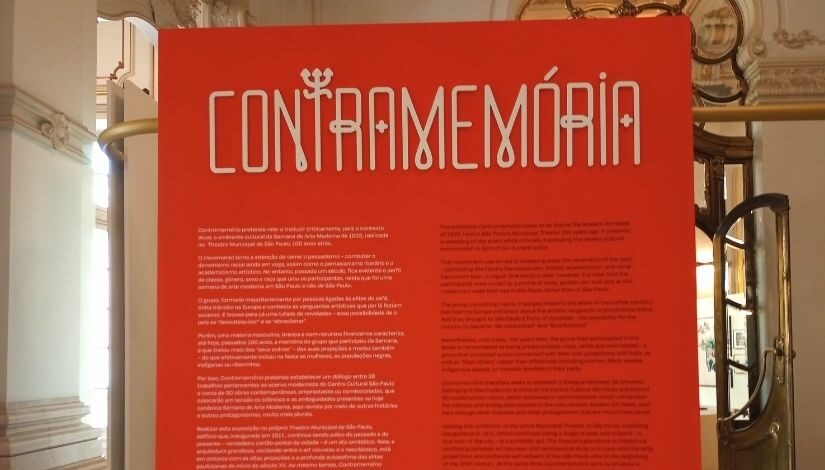 Capa da exposição. Está escrito "Contramemória" e há textos que estão desfocados em um fundo de cor laranja.
