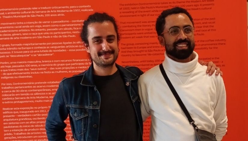 Curadores da exposição em um fundo laranja. O da direita é o Bruno Imparato, veste camiseta preta e jaqueta jeans. Ao esquerda Jaime Lauriano, veste camisa branca de gola alta e usa óculos.