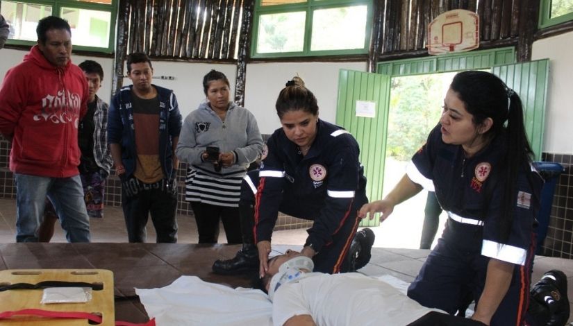 Fotografia de duas socorristas fazendo demonstração de primeiros socorros em uma pessoa que está deitada no chão, ao redor delas outras cinco pessoas observam.