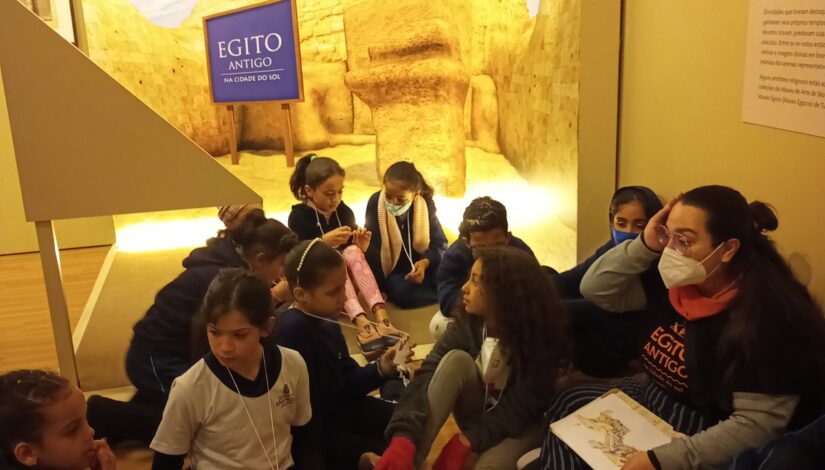 Grupo de crianças sentadas no chão com uma das educadoras da exposição. Ao fundo vemos a figura da Esfinge com a placa "Egito Antigo: Na Cidade do Sol"