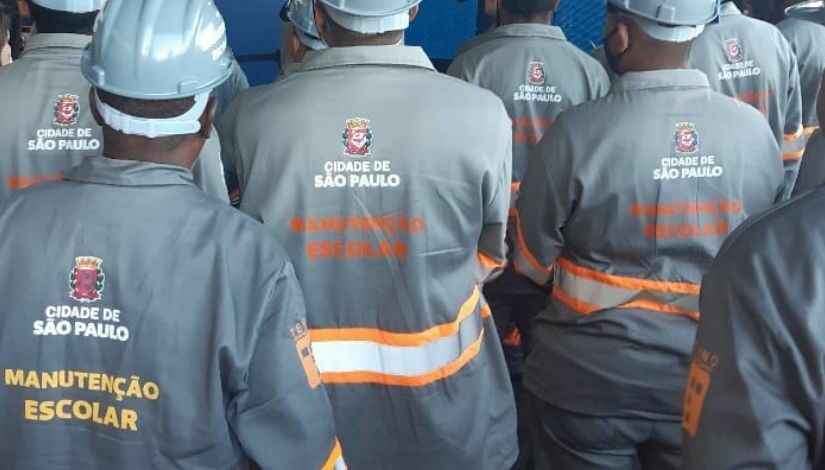 Fotografia mostra vários homens de costas com capacete e usando uniforme cinza com a logomarca 'Cidade de São Paulo' e o texto em amarelo e laranja 'Manutenção Escolar'.
