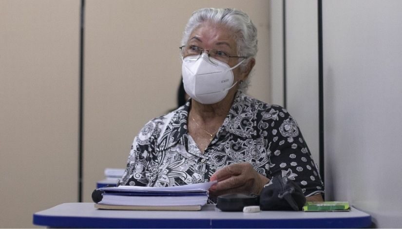 Fotografia de uma estudante da Educação de Jovens e Adultos. Ela possui cabelos grisalhos, usa óculos e máscara de proteção individual. Ela está sentada atrás da sua carteira escolar e sobre a mesa tem seu material escolar.