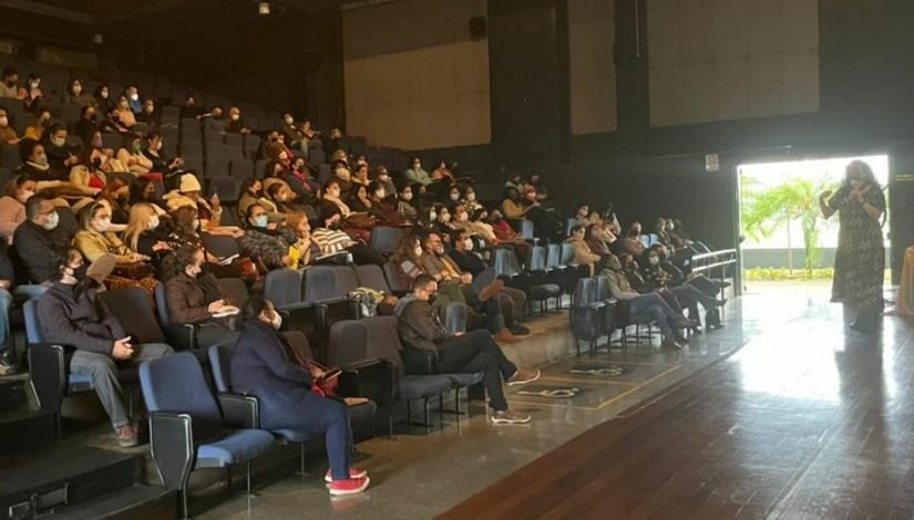 Auditório de um teatro com pessoas assistindo uma palestra. No palco uma mulher conversa com a plateia.