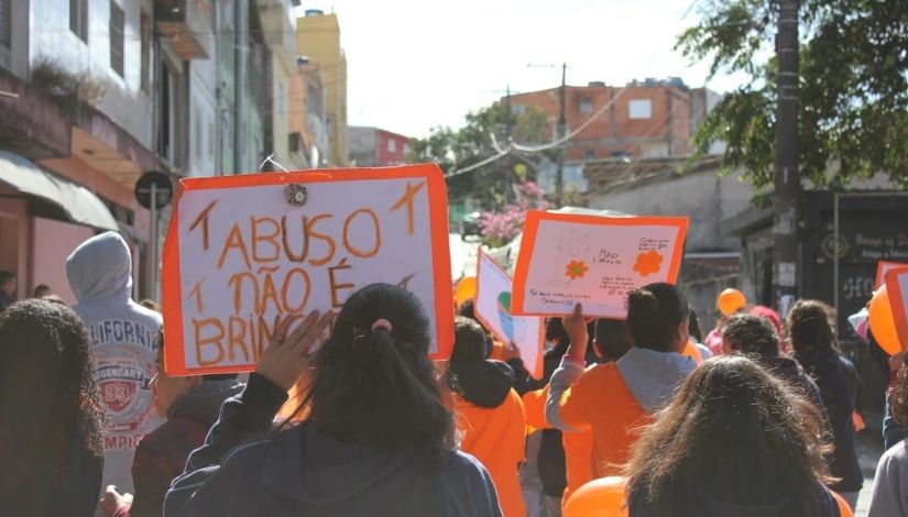 Fotografia de uma passeata com cartazes nas cores branco e laranja. Em um dos cartazes está escrito "Abuso não é brincadeira". As pessoas estão de costas para a câmera fotográfica.
