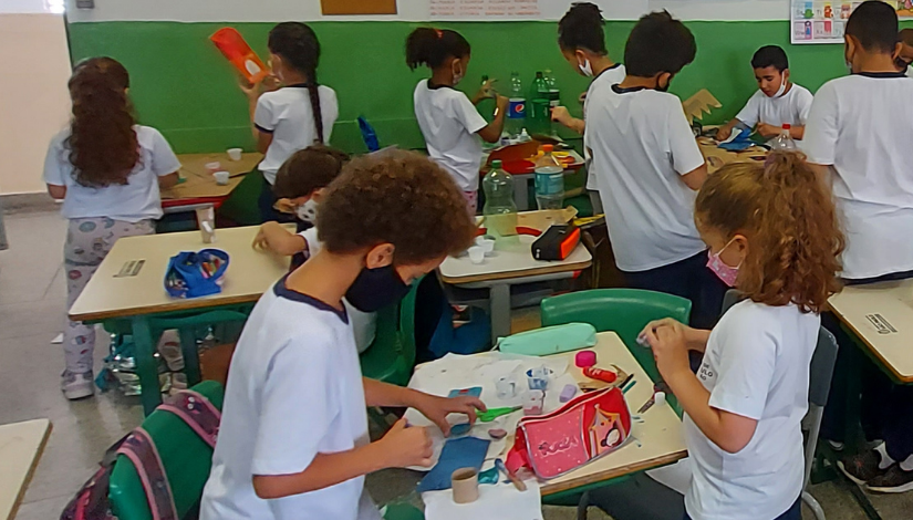 Fotografia de uma sala de aula onde os estudantes estão em grupo ao redor de suas mesas e fazem atividades com materiais reciclados como garrafas pet, tampinhas, papeis, entre outros.