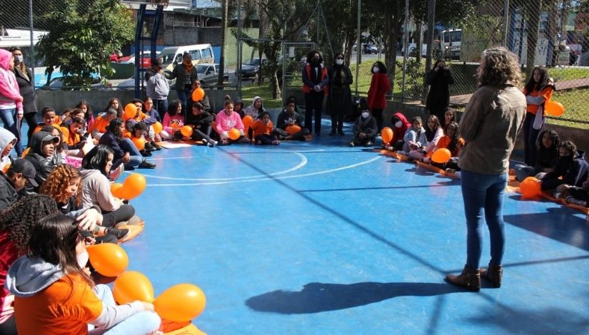 Crianças e adultos sentados em roda na quadra da praça. Alguns seguram balões laranja e usam camisetas da mesa cor.