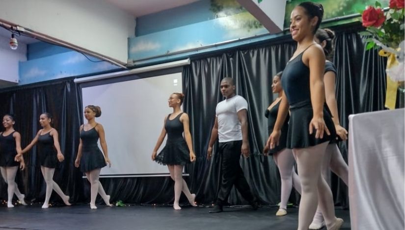 Fotografia de formandos de balé no palco. Mostra 7 bailarinas, usando collant e saia preta, meia calça e sapatilha cor de rosa; e um bailarino de camisa branca e calça preta.