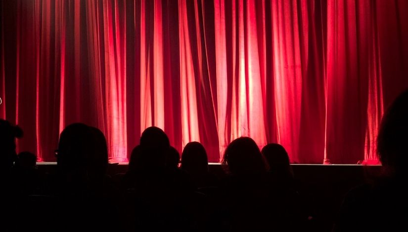 Imagem de uma cortina vermelha do palco com a silhueta de algumas pessoas dos ombros para cima.