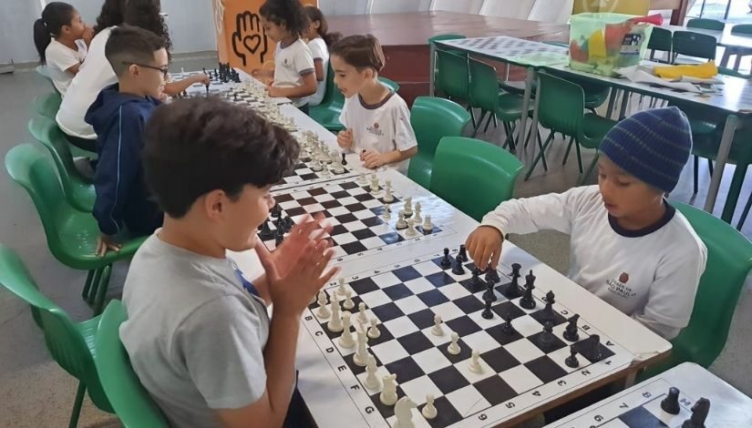 Fotografia mostra estudantes jogando xadrez. O tabuleiro está em cima da mesa e eles estão sentados em volta, cada dupla com seu tabuleiro.