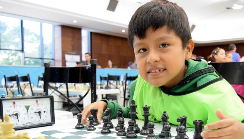 Fotografia em primeiro plano de um menino olhando para lente da câmera fotográfica. Ele está sentado à mesa com um tabuleiro de xadrez sobre ela. A criança está usando blusa e colete verde. Ao fundo outras mesas com tabuleiros de xadrez e outras crianças sentadas.