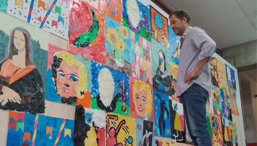 Fotografia de um homem em pé. Ele olha para um painel com obras feitas por estudantes com sacolinhas plásticas que são releituras de pinturas famosas.