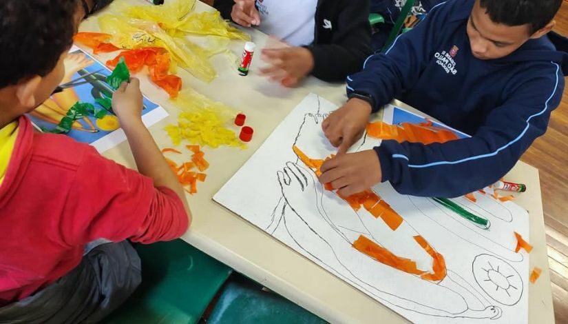 Estudantes sentados em volta de uma mesa produzindo com sacolinhas plásticas uma releitura da obra Abaporu de Tarsila do Amaral.