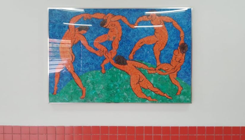 Releitura da obra "A Dança", de Henri Matisse, feita com sacolinhas plásticas por estudantes. O quadro está em uma parede branca com uma faixa de azulejos vermelhos abaixo.