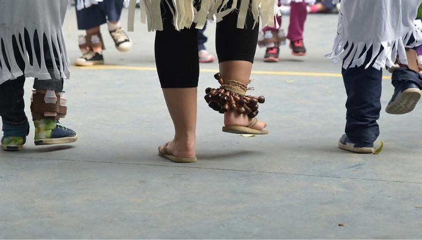 Fotografia fragmentada mostra 3 pares de pés, todos com o pé esquerdo no chão e o direito pouco levantado. No tornozelo direito da pessoa que está no meio há uma tornozeleira para dança de origem indígena, as pessoas que estão nas extremidades têm uma tornozeleira feita de material reciclável.