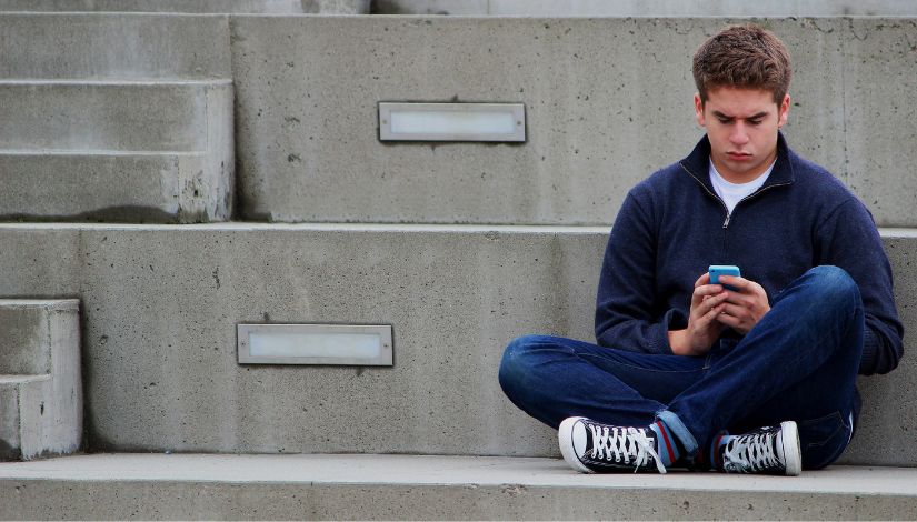 Fotografia de um garoto adolescente sentado em uma escada e mexendo no celular