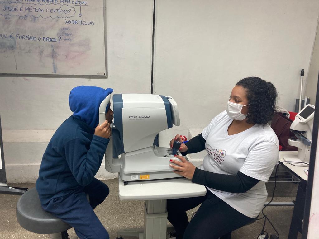 Profissional sentada em frente a um aparelho oftalmológico e do outro lado da máquina um estudante com o rosto apoiado no aparelho.