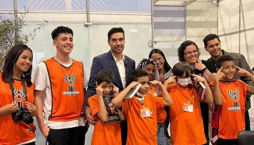 Fotografia mostra 11 pessoas. Da esquerda para a direita, uma menina com colete laranja do Imprensa Jovem e uma câmera fotográfica no pescoço; um menino com o mesmo colete; Abel Ferreira, técnico do Palmeiras, está de paletó azul escuro e camisa social branca; ele abraça um menino que está de camiseta laranja, segurando um livro e com os dois dedos indicadores em sua testa; ao lado direito do menino tem mais três meninos que estão com mesma roupa e pose. Atrás deles há duas meninas, uma mulher e um homem; só aparecem seus rostos.