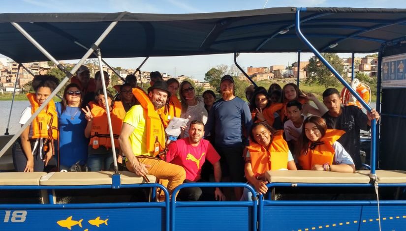 Adolescentes e adultos em um barco azul posando para a fotografia. A maioria usa coletes salva-vidas cor de laranja